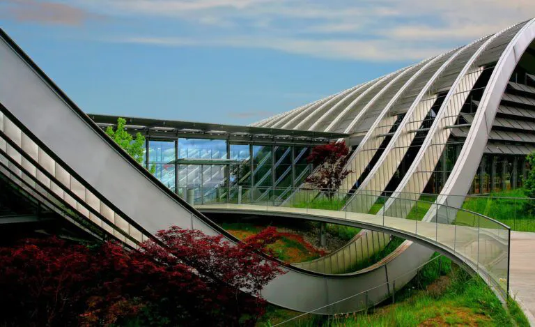 Modern architectural structure with curved steel and glass elements, integrated into a natural landscape with greenery and red foliage, highlighting sustainable and innovative design.